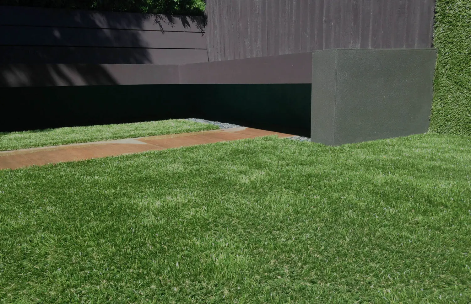 A neatly trimmed green lawn, possibly from a recent artificial grass installation, lines a narrow concrete path beside a modern, dark-colored wall and fence. The scene in El Mirage is well-lit, clean, and minimalist.