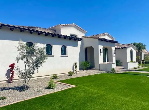 A modern, single-story white stucco house with arched windows and a tile roof sits under a clear blue sky, surrounded by manicured synthetic turf and desert landscaping.