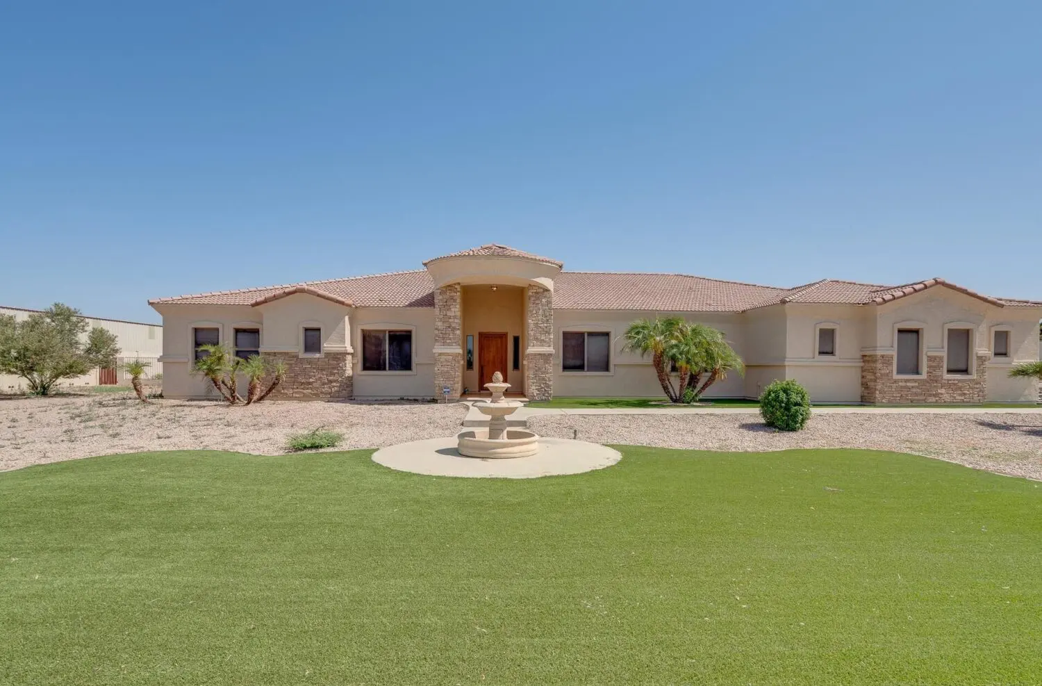 A single-story beige house with stone accents, a red-tiled roof, and a small front porch. A circular fountain sits on a lawn featuring artificial grass installation bordered by desert landscaping and palm plants under the El Mirage sky.