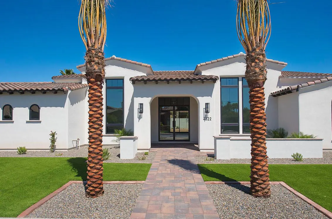 Modern white stucco house with a tile roof, large windows, and two tall palm trees in front. A paved walkway leads to the entrance, surrounded by green grass and gravel landscaping featuring expert artificial turf installation.