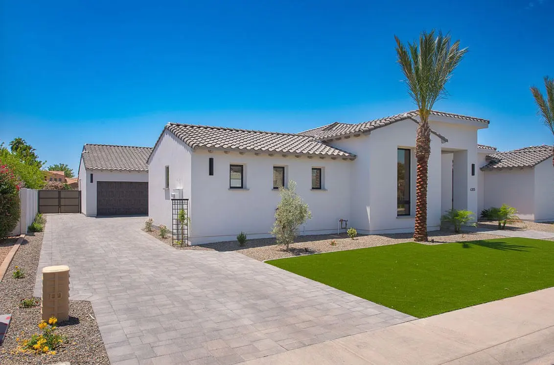 Modern single-story white house with a gray tiled roof, attached garage, manicured front lawn with synthetic turf, a palm tree, and a stone driveway under a clear blue sky.