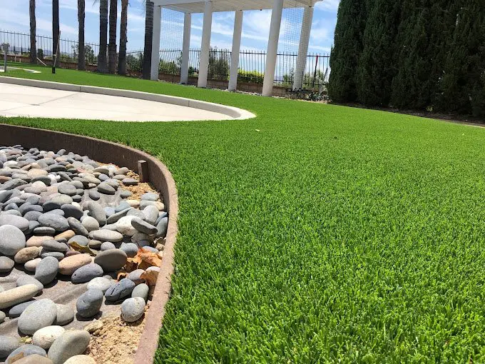 A close-up view of a landscaped yard featuring vibrant artificial turf lawn, a curved metal edge, smooth decorative river rocks, and a white pergola with tall trees and fencing in the background under a blue sky.