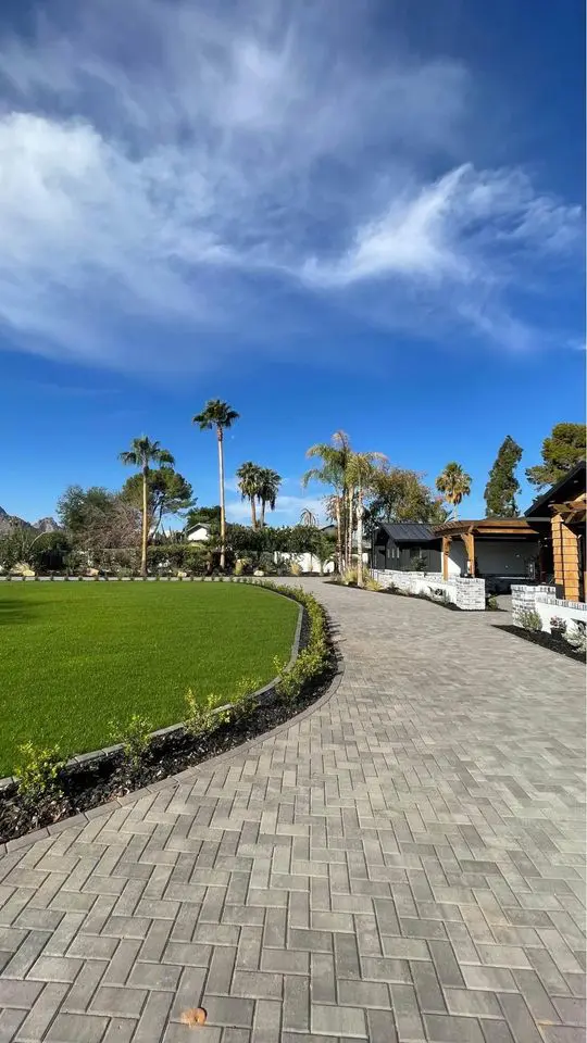 A paved walkway curves through a landscaped area with green grass, palm trees, and shrubs under a bright blue sky with scattered clouds, enhanced by an artificial turf lawn next to modern buildings.