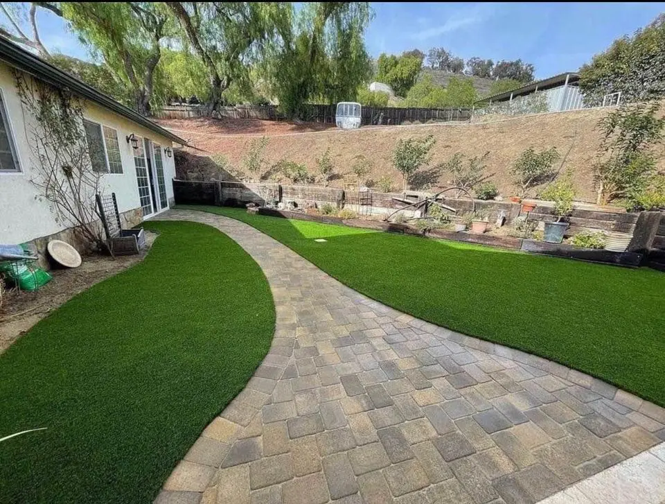 A backyard in El Mirage with a curved stone pathway dividing two sections of lush artificial grass, bordered by a house on the left and a raised dirt garden area with small plants and trees—perfectly crafted by expert artificial turf installers.