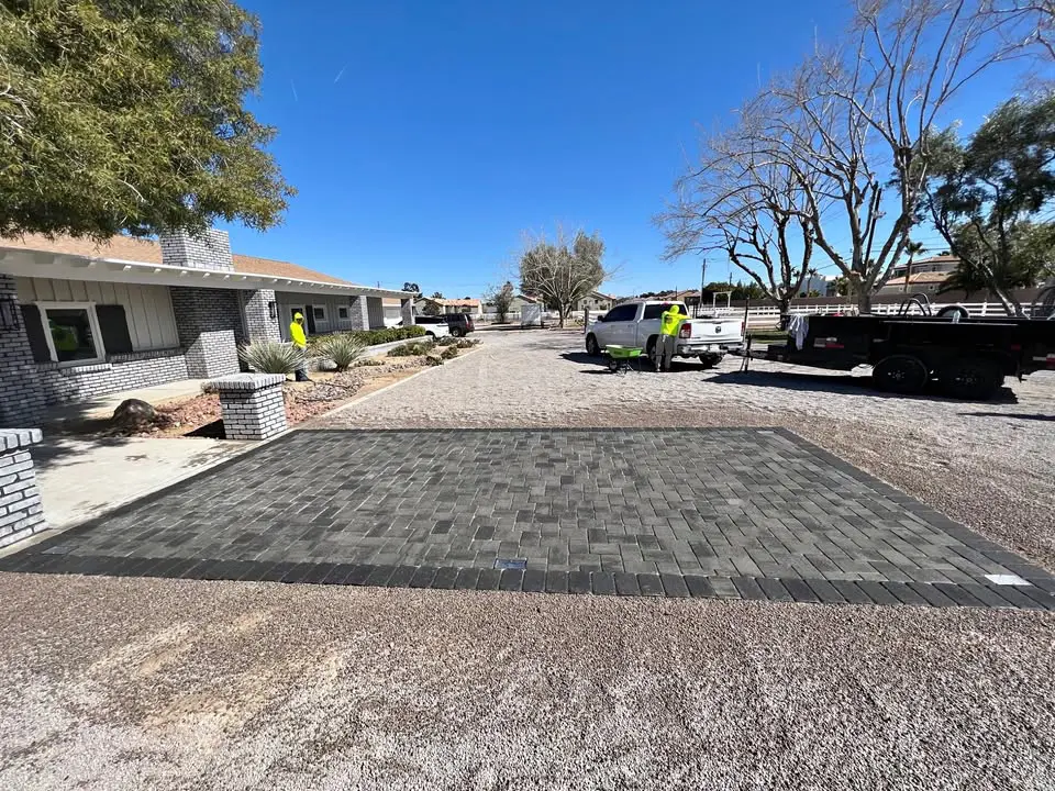 A newly installed rectangular section of dark pavers, bordered by gravel, sits in front of a house in Tolleson AZ. Two workers and utility trucks are on the driveway under a clear blue sky, with leafless and leafy trees nearby.