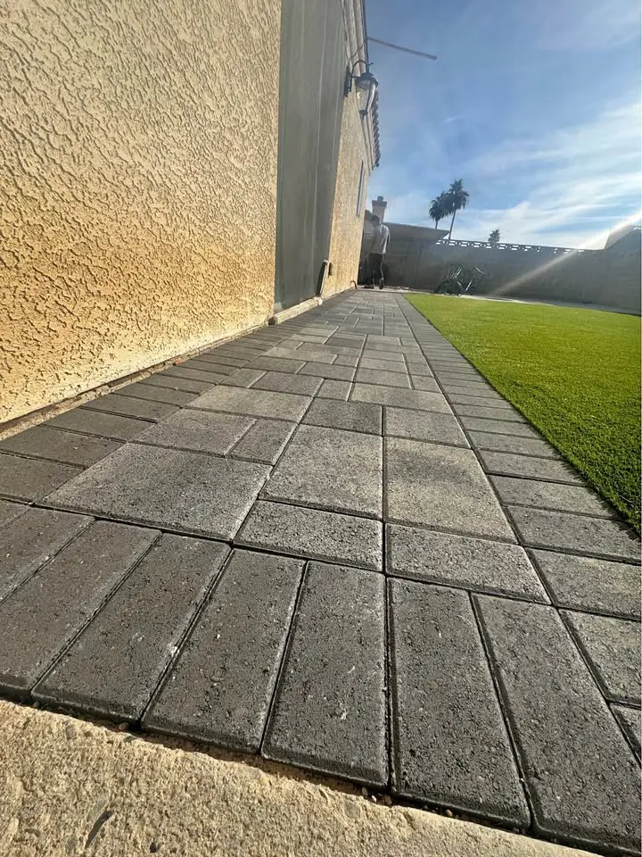 A paved stone walkway runs alongside a beige stucco house, bordered by a neatly manicured artificial turf lawn. The sky is clear and blue with sunlight casting shadows on the ground, highlighting the quality artificial grass installation.