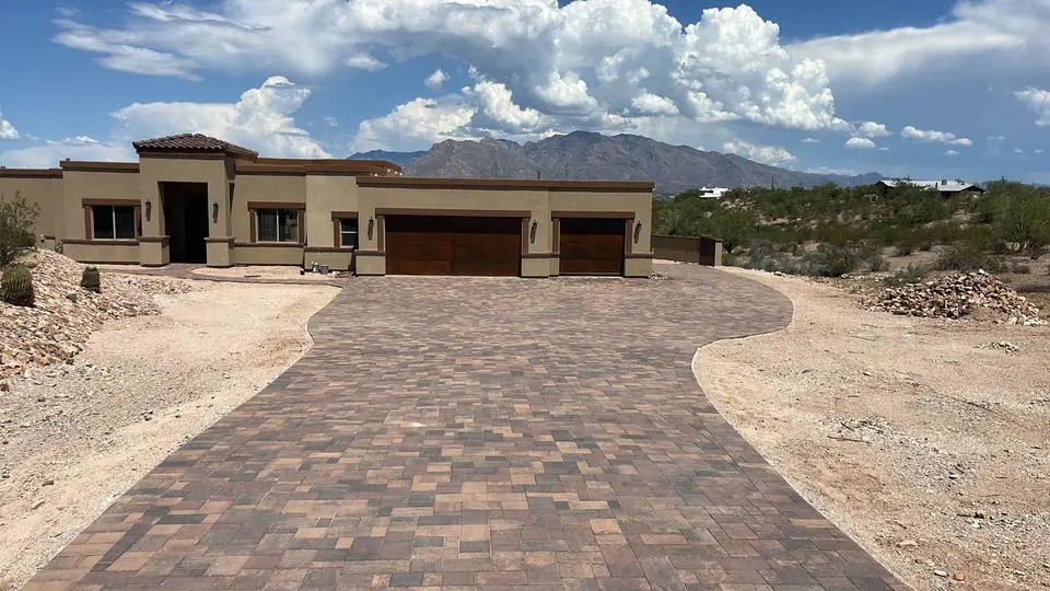 A modern single-story house with a large stone driveway and artificial turf, surrounded by desert landscape and mountains in the background under a partly cloudy sky.