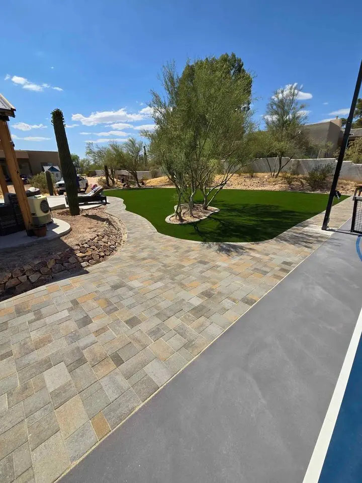 A backyard with a stone-paved walkway, artificial grass installation, desert landscaping featuring rocks, cacti, and small trees—all set under a clear blue sky.