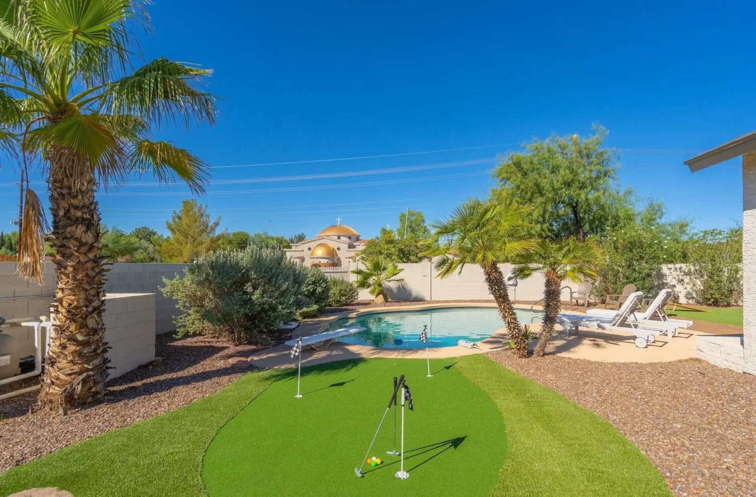 A backyard with a swimming pool, lounge chairs, palm trees, desert landscaping, and a small putting green featuring professional artificial grass installation under a clear blue sky.