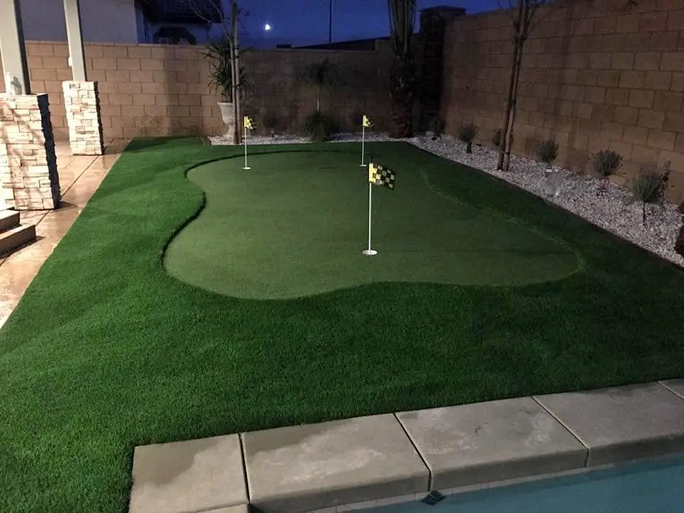 A backyard putting green with three small golf flags, surrounded by neatly trimmed synthetic grass, white rocks, and a brick wall, next to a swimming pool at dusk.