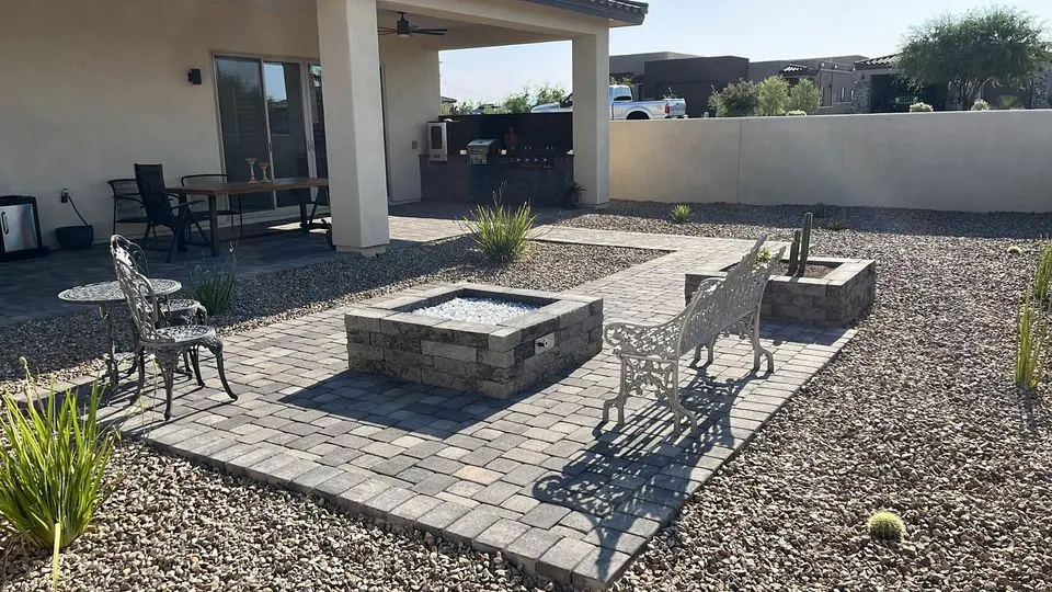 A backyard patio features metal chairs and a bench around a square stone fire pit on pavers, surrounded by gravel, desert plants, and an artificial turf lawn under a covered area next to the house.