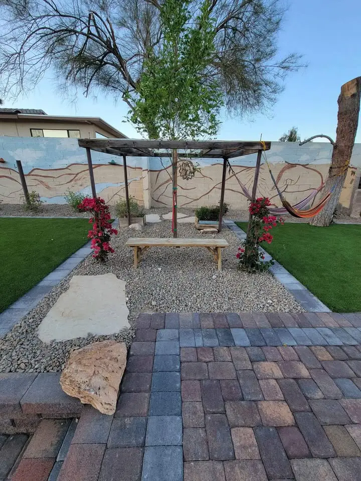 A backyard garden with a wooden pergola over a bench, surrounded by flowering plants and gravel; pavers lead to the area, with synthetic grass on both sides and a hammock hanging to the right.
