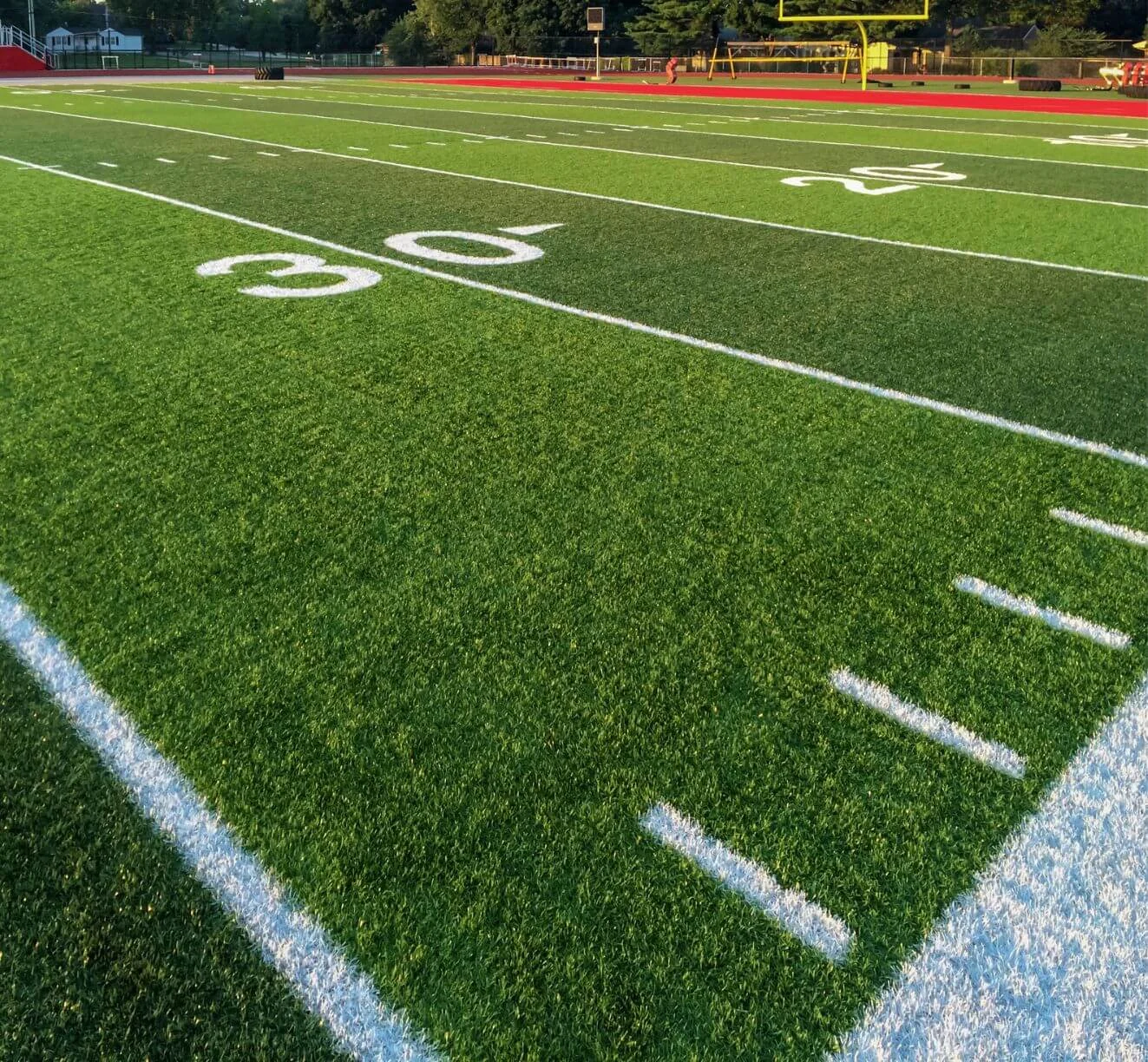 A football field with green synthetic turf, white yard lines, and numbers marking the 20 and 30-yard lines. A yellow goalpost and track are visible in the background under a clear sky.