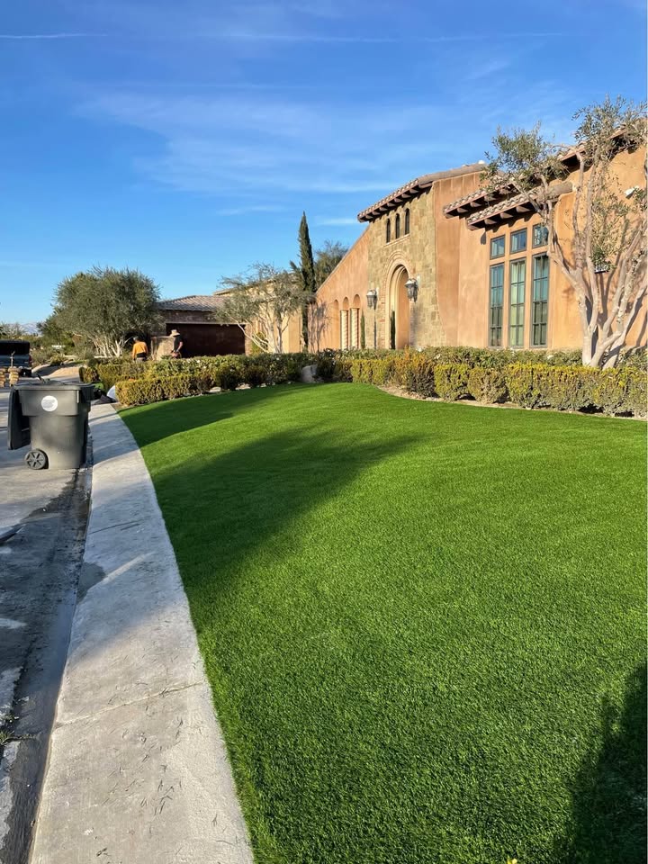 A neatly manicured artificial turf lawn in front of a large, tan stucco house with arched windows and a tiled roof, bordered by trimmed hedges and trees under a clear blue sky. Trash bins are on the curb.
