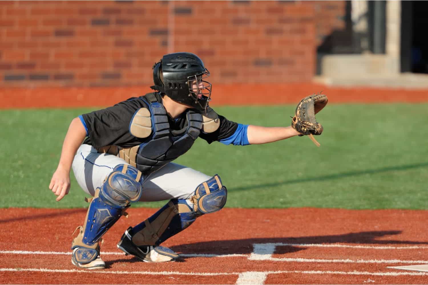 A baseball catcher in full gear is squatting behind home plate on a field with an artificial turf lawn, wearing a helmet, chest protector, and shin guards, with a glove outstretched to catch a pitch.